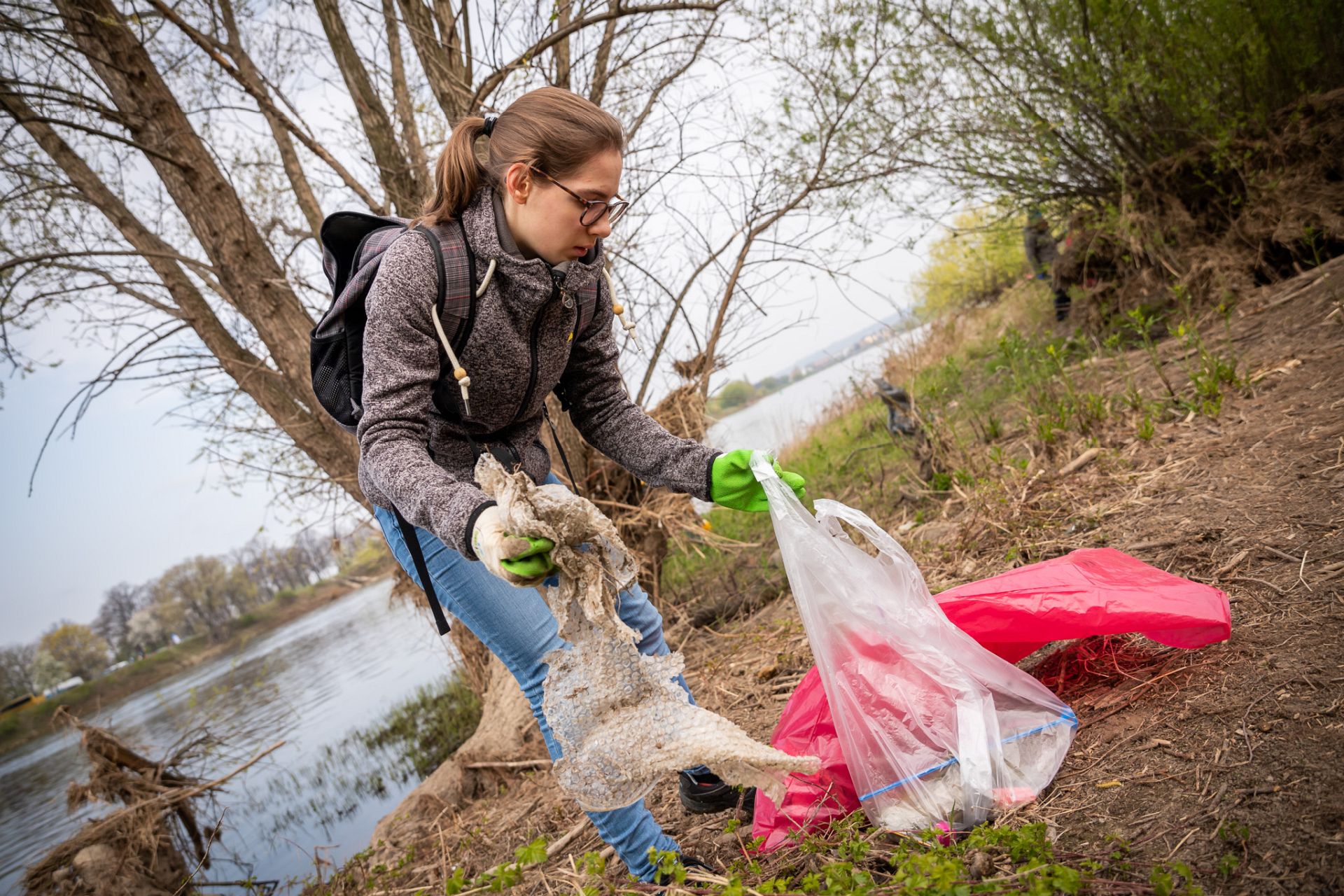 Eine NAJU-Aktive sammelt Müll am Ufer eines Flusses. Sie hält eine große Plastikplane in der Hand und füllt sie in ihren Müllsack.